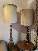 Two vintage tall table lamps side by side on wood surfaces, showing green and gold and black and gold carved spindle bases with tall off-white fabric shades.