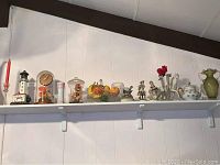 Wide shot of shelf with lighthouse mantle clock, candlesticks, glass dome, faux fruit bowl, porcelain figurines, and vases arranged in a line.
