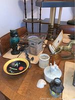 Photo shows a bronze owl sculpture, a small wooden birdhouse, and several decorative items including a white ceramic candle holder and small figurines arranged on a wooden table with a brass table lamp and other items in background.