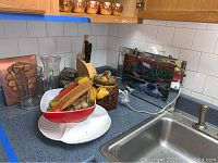 Kitchen counter with vintage Pyrex baking dish, knife block with knives, ceramic cookie jar, owl sculpture, glass pitcher, tiered fruit basket, and various kitchen items.