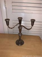 Sterling silver three-armed candelabra standing on a wooden surface in front of white louvered doors.