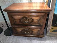 Front view of one nightstand showing two drawers with carved decorative faces and metal ring pull handles, medium brown wood finish.