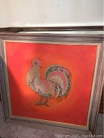 Framed art piece featuring a rooster rendered with metallic texture on an orange background in a wide wood frame.