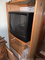 Full view of the wooden entertainment center with glass doors, showing the CRT TV in the center shelf, storage drawers below, and visible signs of wear on the wood surface.