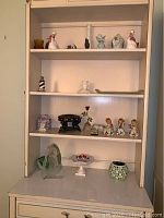 Shelf unit displaying various ceramic and glass figurines, vases, a trinket box, votive holders, and decorative items arranged on multiple shelves.