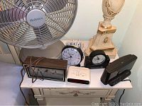 A collection of six table clocks of mixed styles and two table fans placed on a white dresser, all untested.