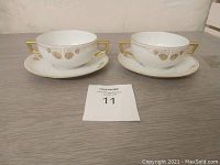 Pair of white porcelain soup cups with gold decorative handles and pattern on plates shown on wooden surface.