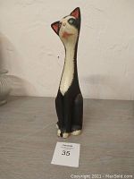 Front view of the tall slender black and white painted wooden cat statue on a wooden surface with textured wall background.