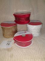Four rolls of Celebrate It brand Christmas ribbons, one in red glitter, one solid red, one gold glitter, and one white glitter, shown grouped in front of a beige background.