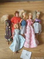 Photo of six dressed dolls including Barbie and similar style dolls on wooden surface with tag 82