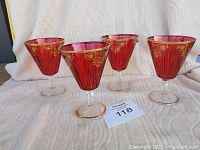 Four red wine glasses displayed showing gold floral detailing on the red bowls and gold-rimmed bases and rims.