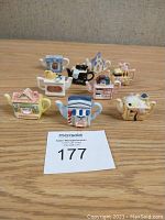 Front view of all 9 miniature teapots arranged on a wooden surface showing various kitchen and house designs in pastel colors