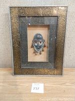 Frontal view of the framed decorative artwork showing a metallic mask with dangling bells mounted on a beige background and surrounded by an ornate golden frame and a textured inner frame.