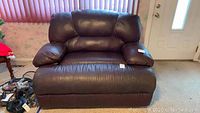 Front view of a large brown leather recliner loveseat placed indoors on carpet, showing cushioned armrests and backrest.