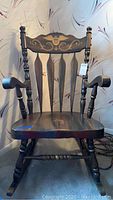 Full view of dark wooden rocking chair showing seat, arms, legs, and rockers resting on floor against light colored wall with floral pattern wallpaper.