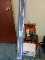 Box containing white curtain rod wrapped in plastic, partially leaning against wall near door