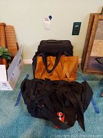 Three bags shown together; one black canvas laptop bag and two tan leather handbags behind it on a carpeted floor.