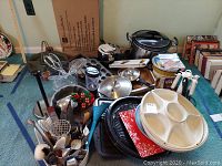 Full assortment of cookware, bakeware, Crockpot, serving trays and utensils laid out on carpeted floor.