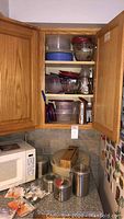 Kitchen cabinet open showing stacked clear and blue plastic food storage containers, glass measuring bowls, stainless steel canisters, and cleaning products on shelves, with a microwave on the counter under the cabinet