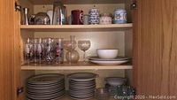 Cabinet shelf showing stacked plates, stacked smaller plates, mugs, and glasses including decorated and plain glassware.