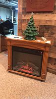 Electric fireplace standing on carpeted floor with glowing ember display visible through glass front panel. Wood finish cabinet with decorative columns.