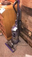 Full view of Dyson Ball Animal 2 upright vacuum cleaner standing on carpet next to wooden furniture showing handle, hose, dust bin, and cord.