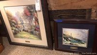 View of both framed prints leaning against wall. Larger print is Victorian Garden II with large house and garden. Smaller print is Clearing Storms with lighthouse scene. Both frames are dark wood with ornate styling.