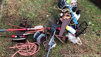 Overall pile of rakes, vise, garden weasel, extension cords, and other assorted hand tools on grass surface