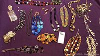 Photo showing variety of necklaces, earrings, and bracelets laid on purple cloth, featuring blue, black, and orange multi-strand beaded jewelry and small clip-on earrings.