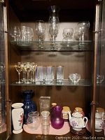 Two shelves inside a wooden cabinet showing various crystal and glass water glasses, wine glasses, and small vases including blue and purple glass ones as well as ceramic pitcher with floral decoration.