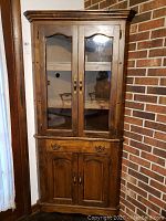 Front view of the corner cabinet showing glass doors with brass handles and the lower cabinet with two wooden doors and a drawer.