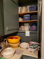 Photo of shelves and cabinet corner showing vintage Tupperware bowls stacked, plastic food storage containers in plastic baskets on shelves, folded kitchen towels, and other kitchen items.