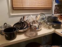 Several pots and pans including a large black pot with lid, multiple frying pans in black and stainless steel finishes with metal handles, a set of nested stainless steel saucepans on a countertop.