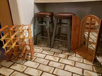 Two wood stools with round brown cushioned seats and gray legs, wooden foldable wine rack, and mirrored decorative shelf displayed on a tiled floor.
