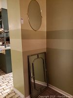 Two wall mirrors: an oval brass/gold-tone framed mirror hanging on the wall, and a larger rectangular mirror with a decorative faceted glass frame leaning against the wall.