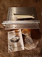 Metal tile saw with yellow base and sliding guide rail viewed from above with instruction manual and some small parts in plastic.