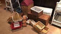 Wide shot showing assortment of wicker baskets including a dollhouse-shaped storage hamper, bread baskets, condiment caddy, letter organizer, and other wicker storage boxes and baskets in various sizes and colors.