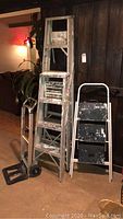 Four-foot ladder, six-foot ladder, small hand truck, and small two-step ladder grouped together indoors.