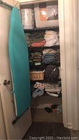 Photo of closet shelving with teal ironing board hung on door, various folded linens, towels, and quilts stacked on shelves, and assorted fabric items showing variety and quantity of linens in lot
