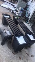 Two long black plastic benches with white planter boxes at each end outside on a concrete surface, near a house wall and utility equipment.