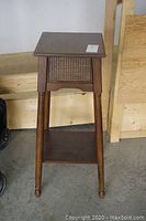 Front view of the wood plant stand showing the square top, woven cane panels beneath the top, four turned legs, and lower shelf.