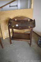 Full view of the wooden butler's tray with brass hinges on a stand showing front and side angles. Surface scratches and wear visible on tray and lower shelf.