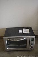 Front angle view of Oster digital toaster oven showing control knobs, display, glass door, handle, rack, and tray with manuals on top.