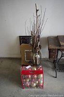 Photo shows wooden framed mirror against wall with vase containing twigs and pine cone decor on metal stand in front. Behind are two picture frames, one burlap style and one HP with damage. Box of Christmas ornaments also visible.