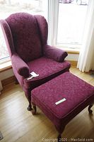 Vintage wingback chair and matching ottoman near window on wooden floor. Chair has burgundy patterned fabric upholstery and claw feet wooden legs.