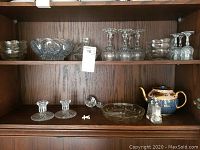 Full view of wooden cabinet shelf holding all glass, crystal, teapot items and pin.