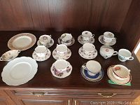 Multiple teacups with matching saucers, two china plates, small teapot, and a small dish laid on a wooden cabinet surface