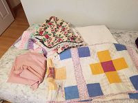 Overview photo showing assorted bedding textiles including the handmade quilt folded, white blanket with hearts, floral Christmas tree skirt, knitted striped blanket, and a single bed sheet all laid out on a bed.