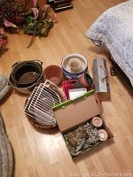 Overview of multiple plant pots, baskets, plant stand, wooden decor items, and empty toolbox on floor near bed.