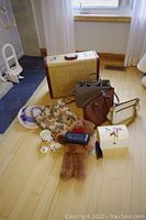Overview photo of vintage suitcase, purses, teddy bear, keepsake box, hat and scarf on a wooden floor near window with curtains